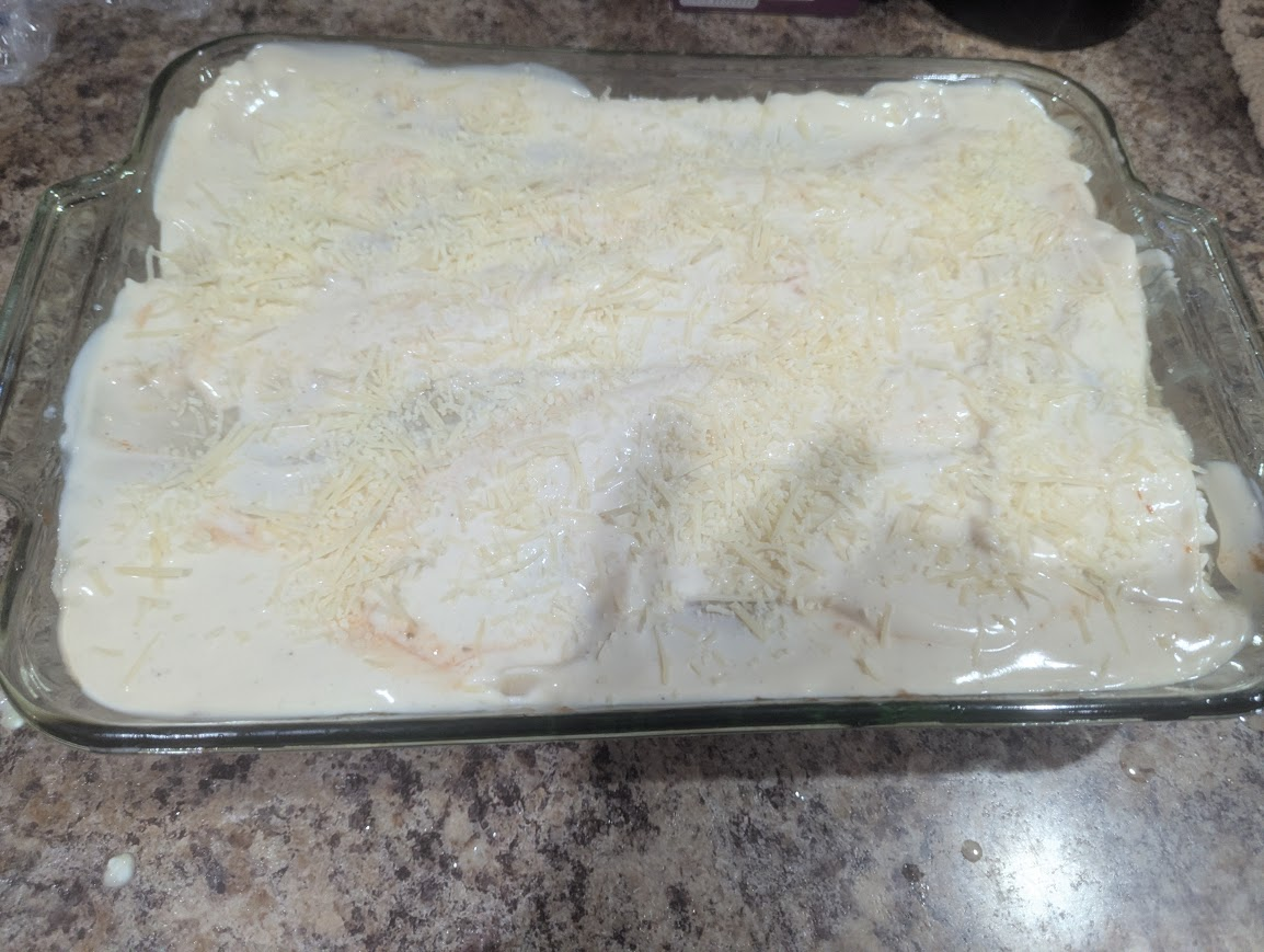 Unbaked Two-Sauce Lasagna topped with Alfredo sauce and Parmesan in a glass dish